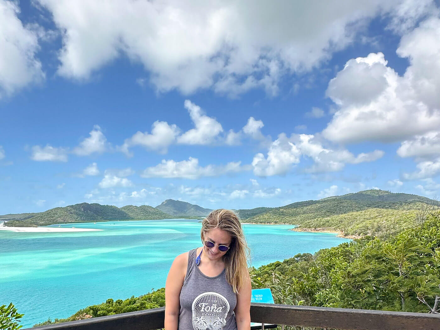 Charlie is standing at the Hill Inlet lookout, looking down with lush green hills and the swirling turquoise and white sands of the Whitsundays in the background under a blue sky with fluffy white clouds.