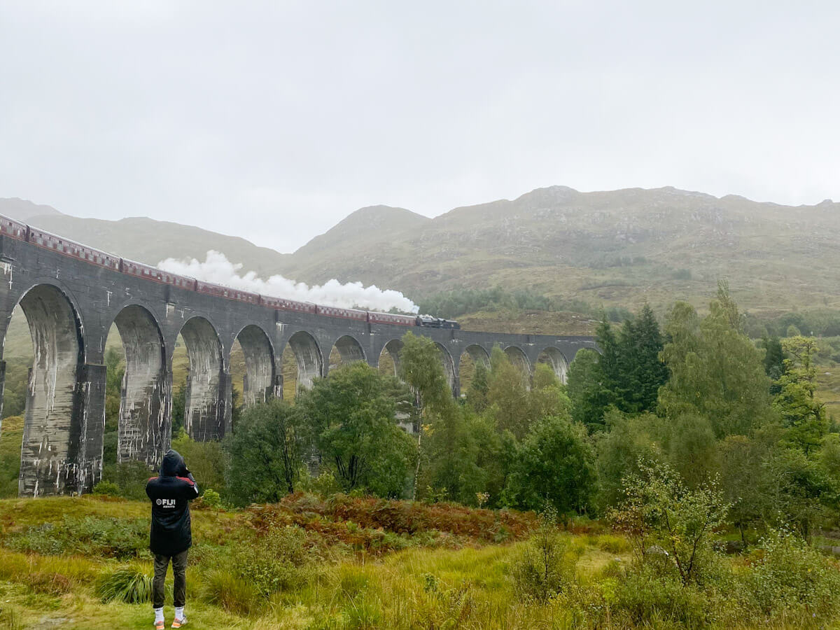 A photographer at the glenfinnan viaduct viewpoint capturing the Jacobite steam train crossing the arches against a backdrop of Scottish hills.