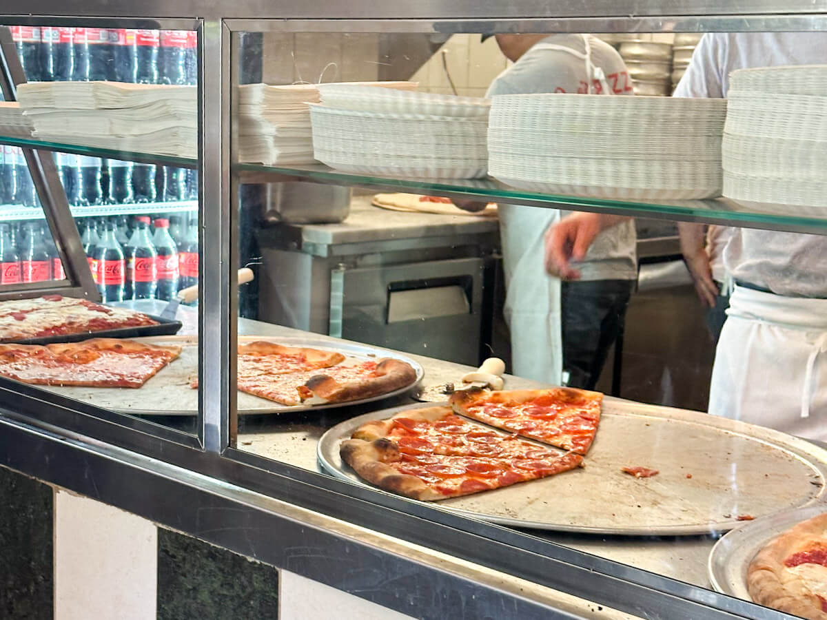 Fresh pepperoni and cheese pizza slices resting on metal trays behind a glass counter at Joe's Pizza in Greenwich Village, highlighting where to find the best pizza in new york as part of your food in new york guide.