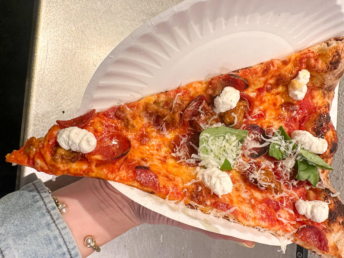 A person holding a paper plate with a slice of L'Industrie Pizzeria pizza topped with cheese, pepperoni, dollops of burrata, and fresh basil, showcasing some of the best pizza in new york as part of the local food in new york scene.