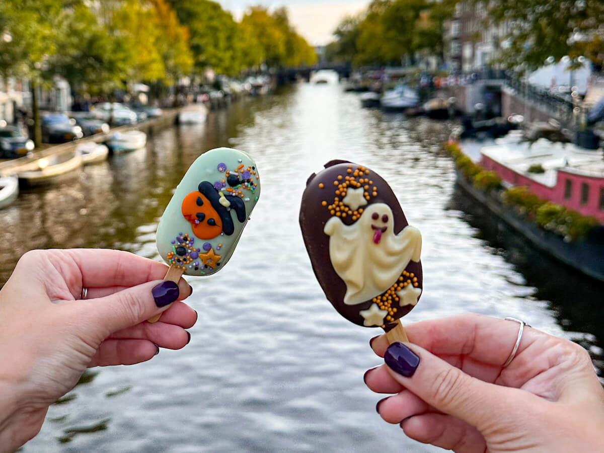 Two hands hold up Halloween-themed cake pops on sticks, featuring spooky decorations against the backdrop of an Amsterdam canal. Making amsterdam one of the best places to travel to in autumn