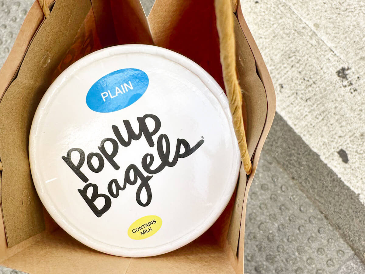 A round container from Popup Bagels labeled "Plain" resting inside a brown paper bag, showcasing popular food in New York.
