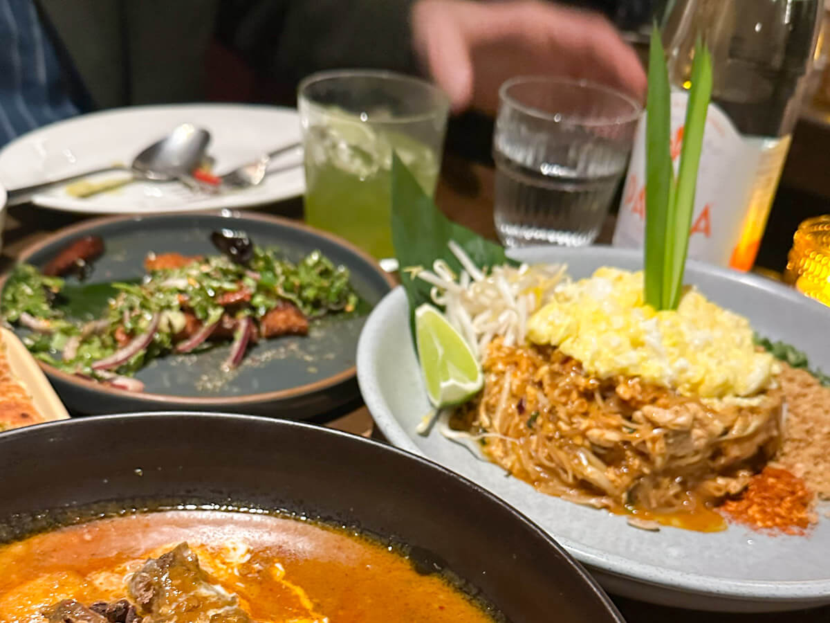 A spread of Thai dishes at Pranakhon featuring pad Thai topped with scrambled eggs and fresh herbs, a bowl of rich curry, and a plate of salad, highlighting the diverse culinary scene for food in new york.