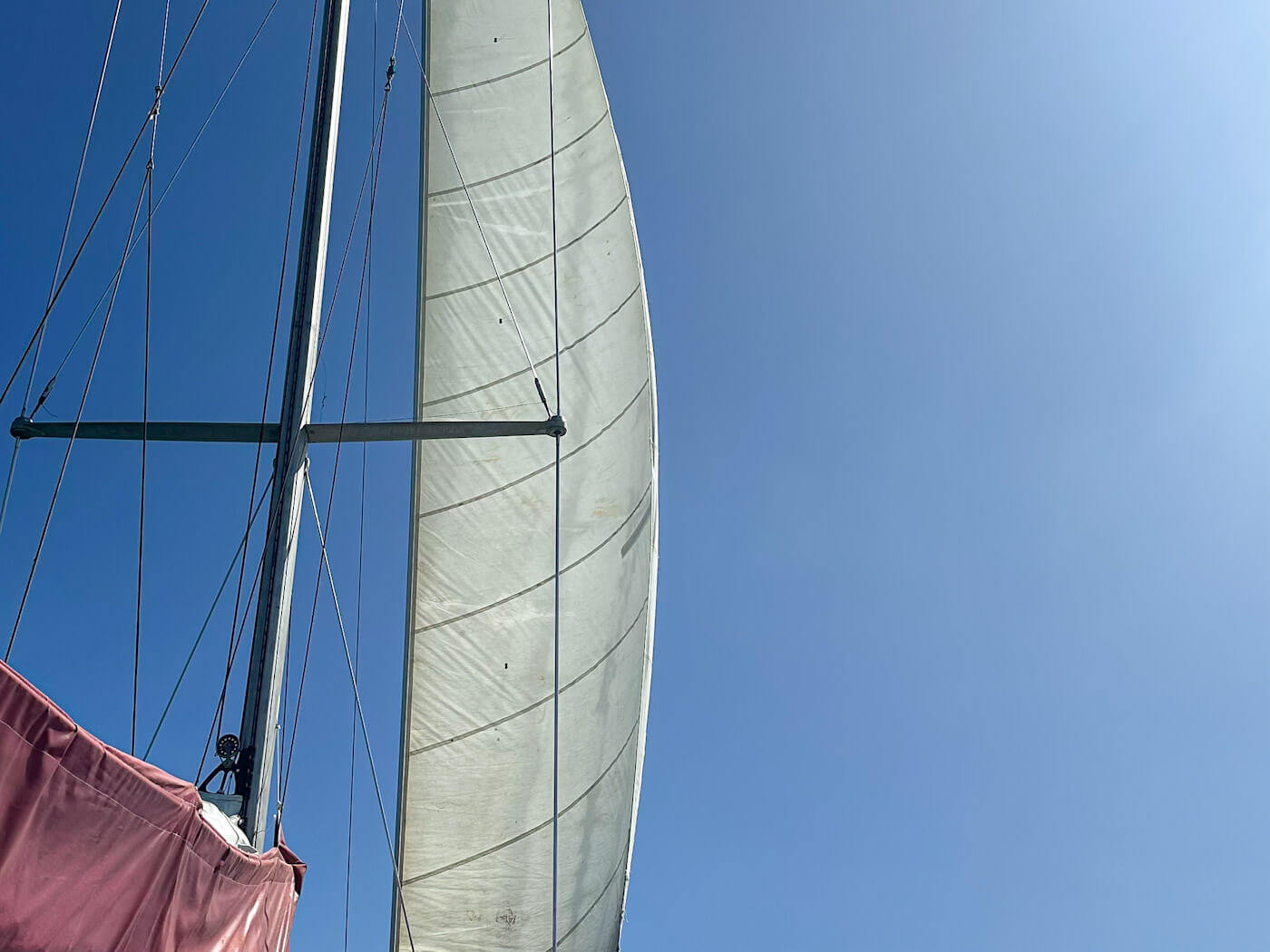 A low-angle shot looking up at a taut white mainsail catching the wind against a clear blue sky, captured while sailing the Whitsundays.