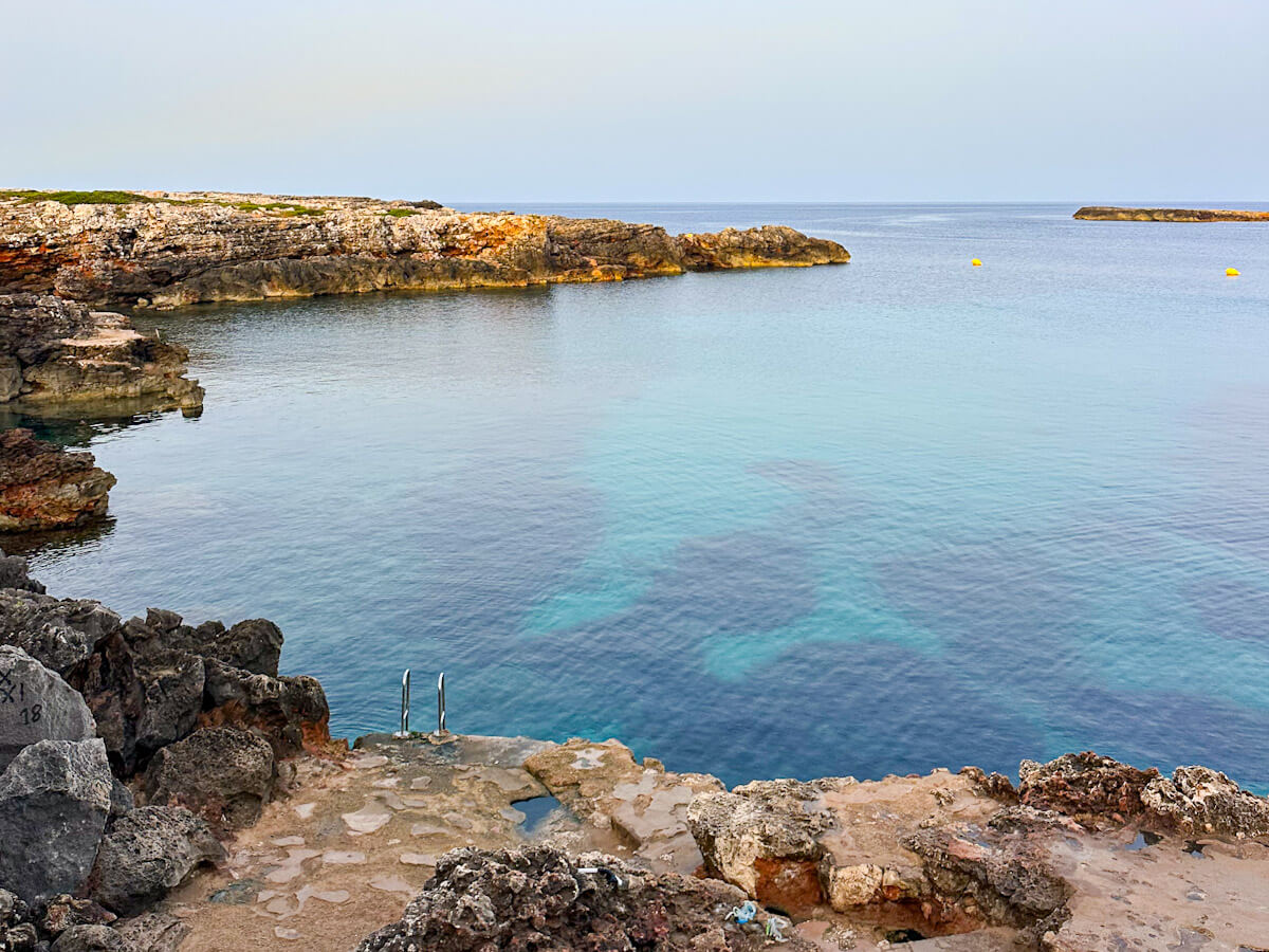 Scenic view of the rocky coast and crystal-clear blue waters at Ses Olles in Menorca, featuring a set of metal swimming ladders built into the rock ledge.