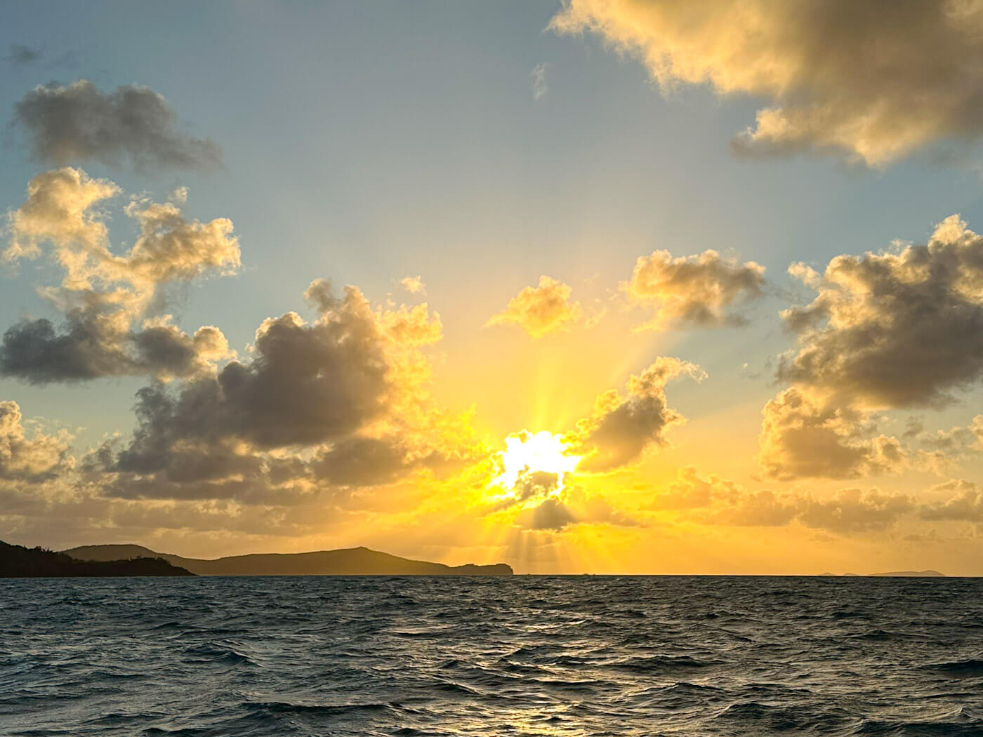 A golden sunrise casting bright sunbeams through scattered clouds over the dark silhouette of distant islands and rippling ocean waters while sailing the Whitsundays.