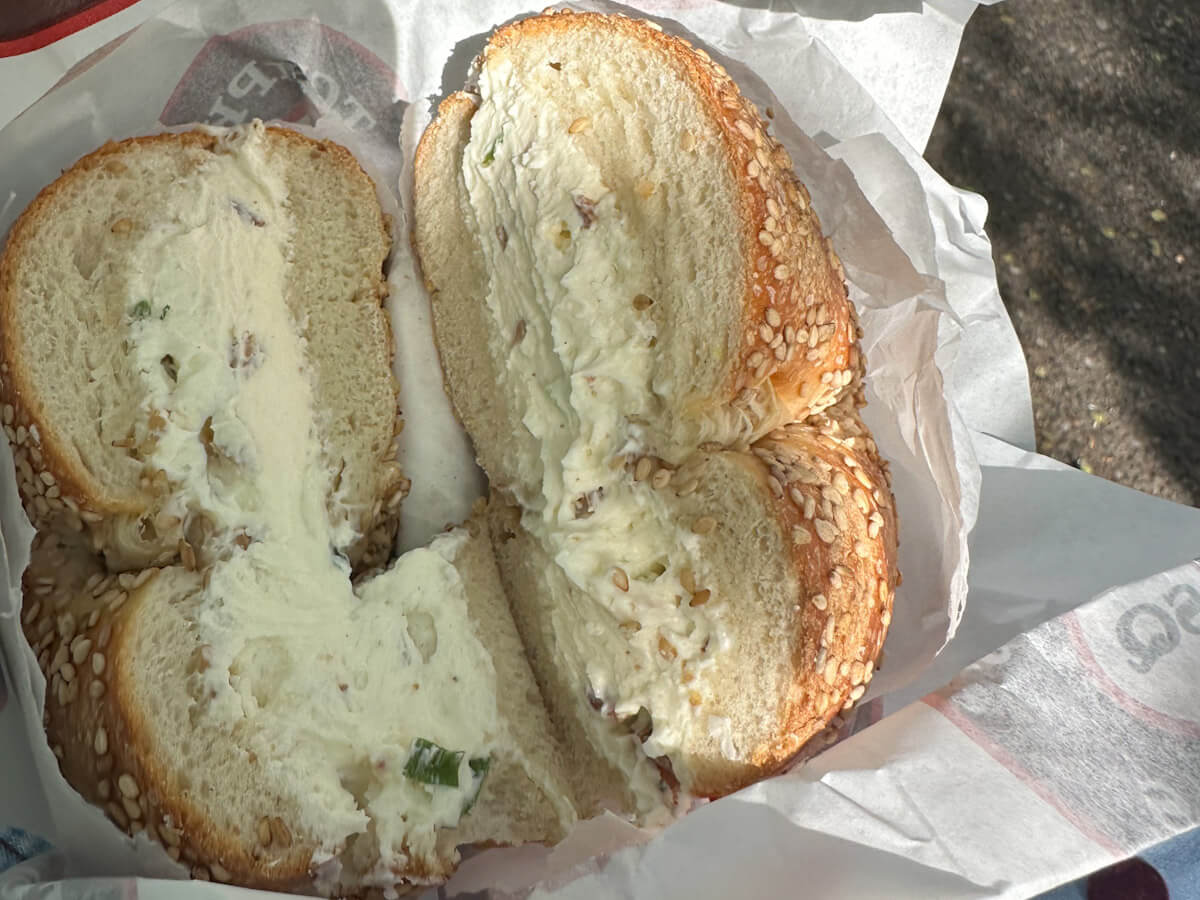 A close-up shot of a freshly sliced sesame seed bagel heavily stuffed with thick cream cheese spread, sitting inside white deli paper, highlighting Tompkins Square Bagels as some of the best bagels in New York.