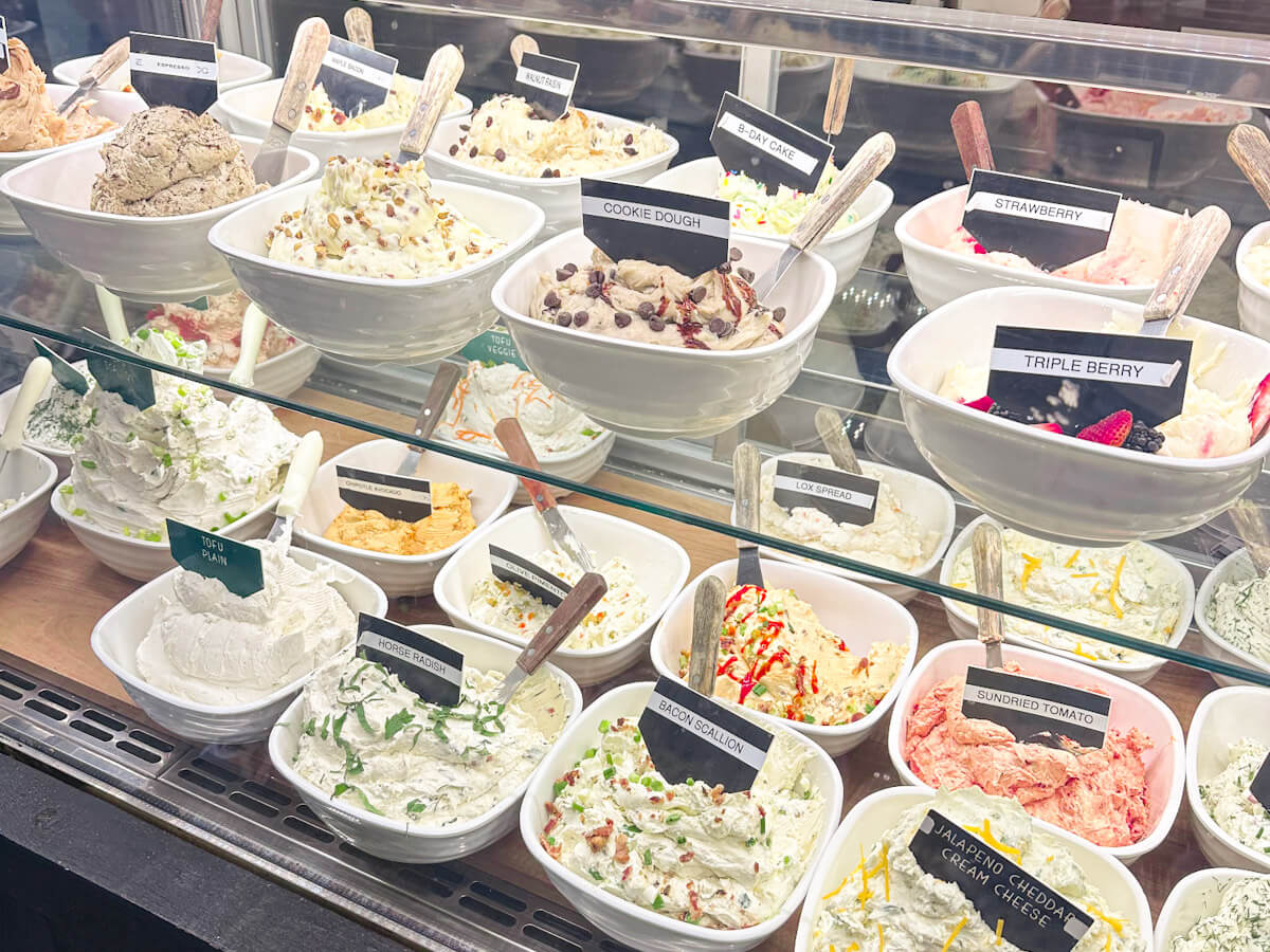 A glass display case filled with rows of white bowls containing various flavored cream cheeses and spreads, each labeled with names like bacon scallion and cookie dough, showcasing options at Tompkins Square Bagels for the best bagels in New York.