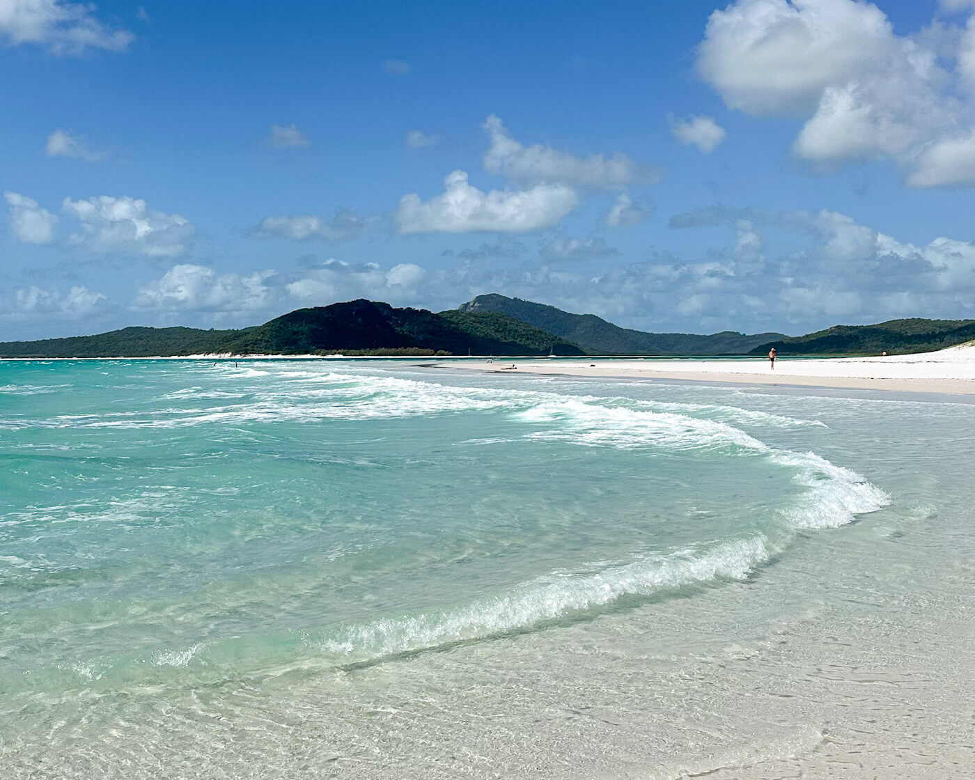 A gentle turquoise wave rolling onto the pristine white silica sand of Whitehaven Beach, with lush green islands in the distance under a bright blue sky dotted with white clouds during a trip sailing the Whitsundays.