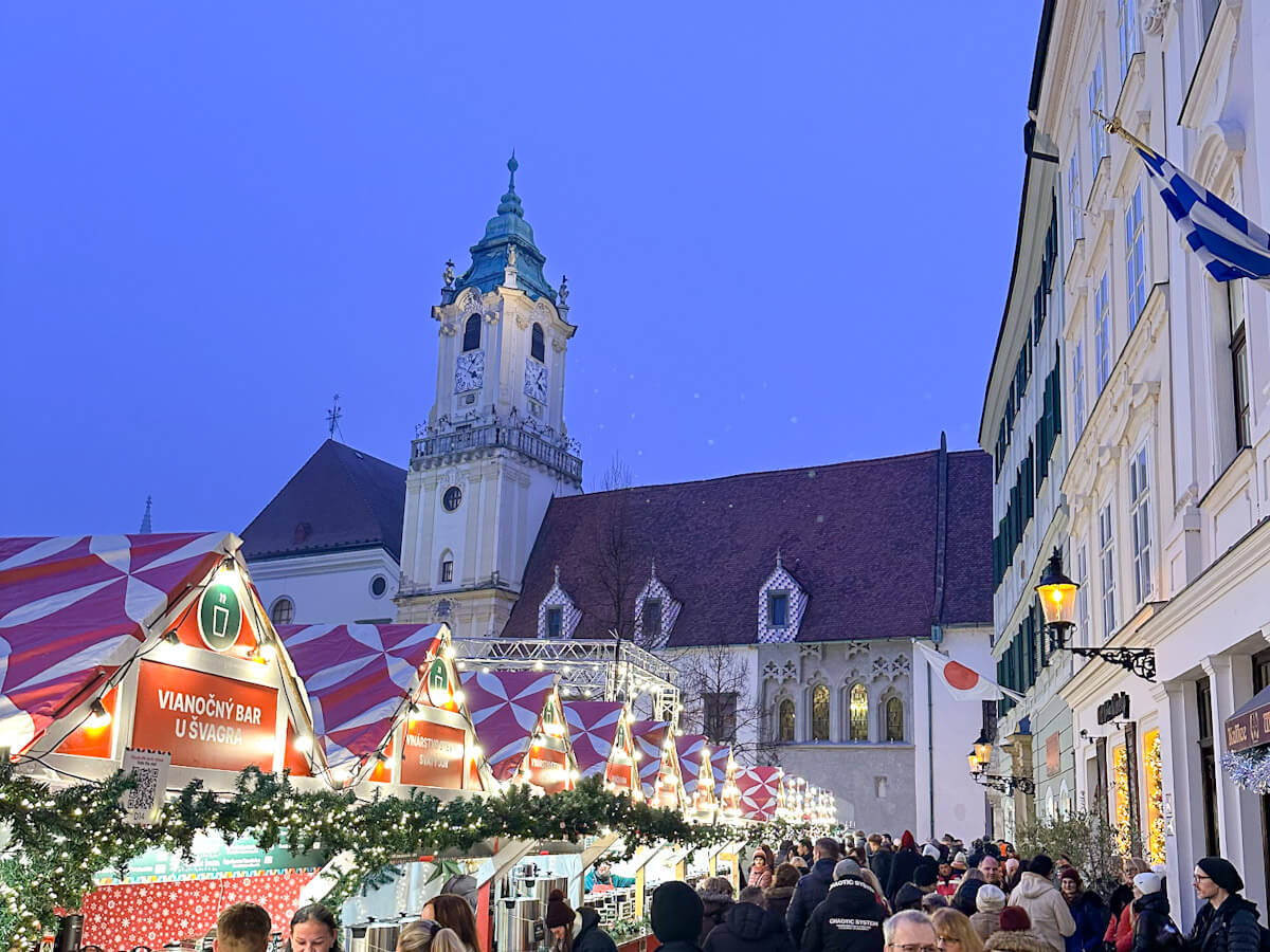 Festive evening view of the Bratislava Christmas Market with illuminated market stalls and crowds under a blue twilight sky, featuring the historic Old Town Hall tower and classic European architecture in the background.