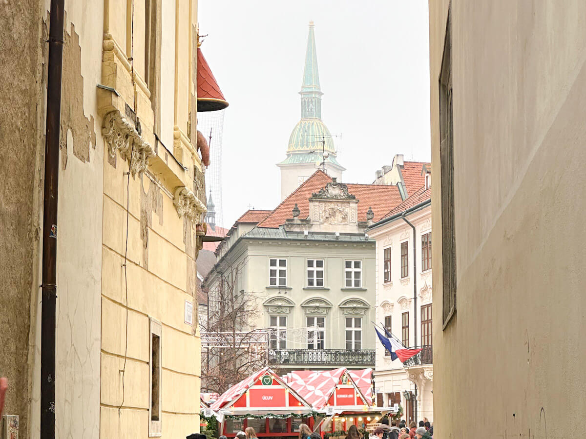 A view framed by historic buildings looking toward the festive stalls of the Bratislava Christmas Market in Frantiลกkรกnske nรกmestie, with the distinctive green spire of the Old Town Hall rising in the background.