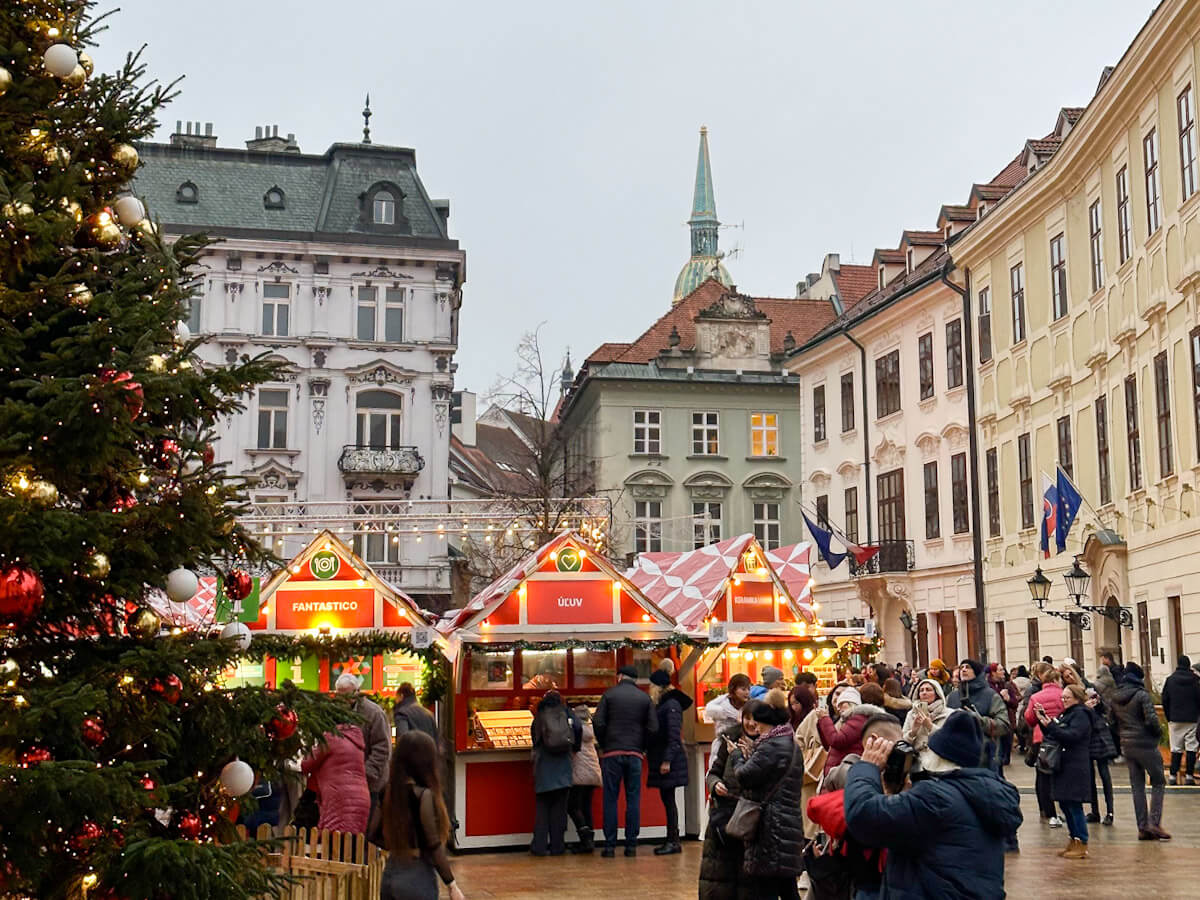 A large decorated Christmas tree beside festive market stalls and crowds of people enjoying Hviezdoslavovo nรกmestie during the Bratislava Christmas Market, framed by historic European buildings and a distant church spire.