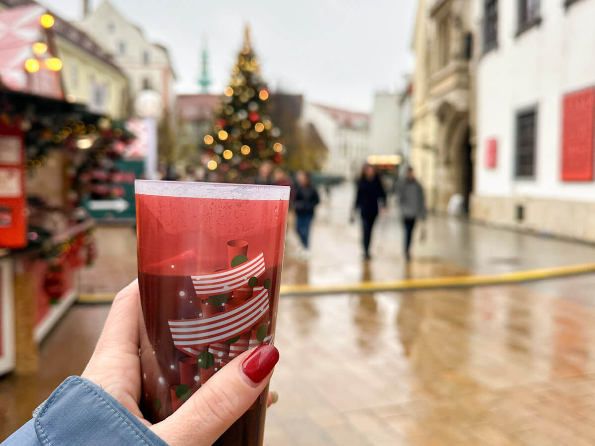 Holding a cup of mulled wine at the Bratislava Christmas Market in Hlavnรฉ Nรกmestie, with festive market stalls, a decorated Christmas tree, and historic square buildings blurred in the background.