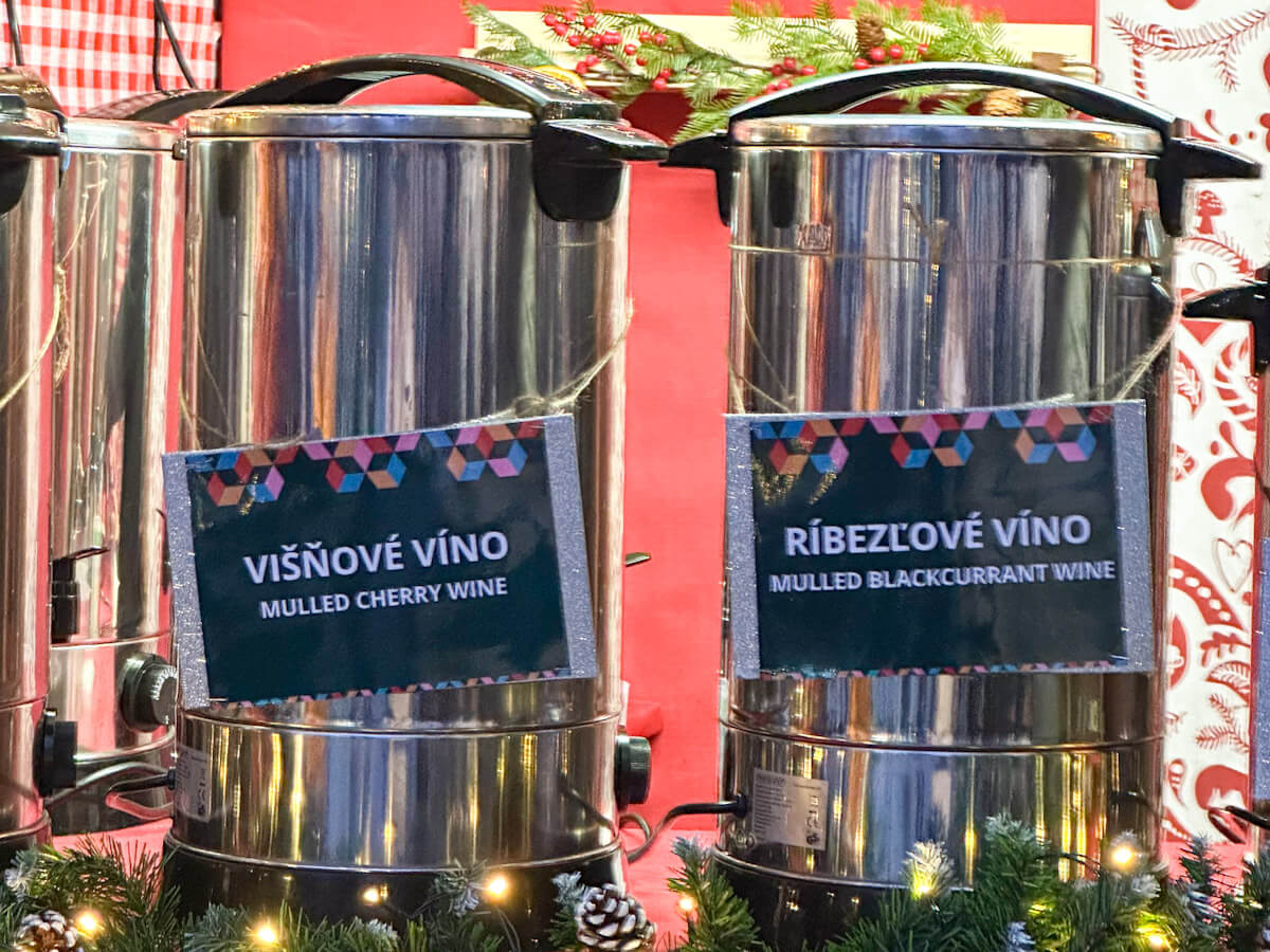 Stainless steel beverage dispensers at a stall during the Bratislava Christmas Market, labeled for mulled cherry wine (viลกลovรฉ vรญno) and mulled blackcurrant wine (rรญbezฤพovรฉ vรญno).
