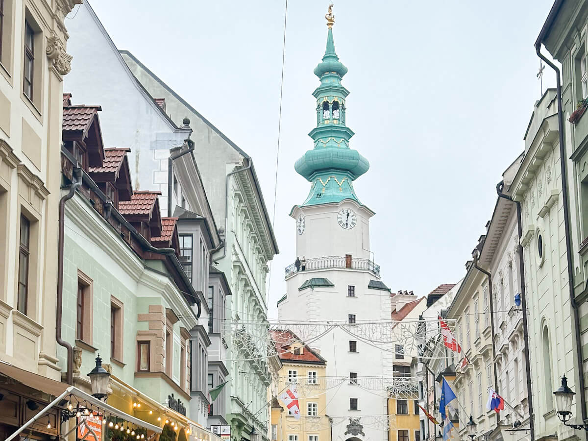 A narrow historic street in the Old Town of Bratislava, Slovakia, framed by colourful buildings and looking toward the distinctive green spire and clock tower of the Old Town Hall.