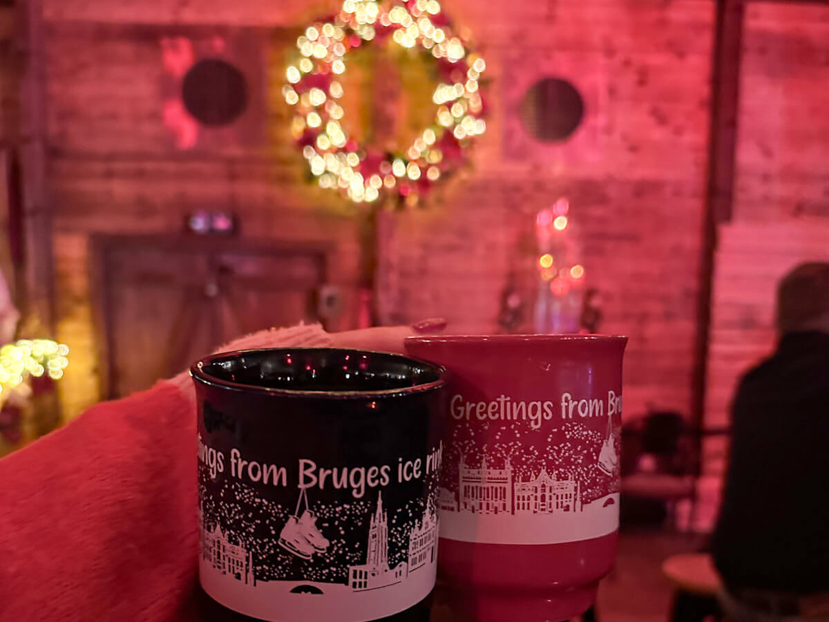 Two mugs reading "Greetings from Bruges ice rink" held up inside the cosy winter bar during Bruges at Christmas