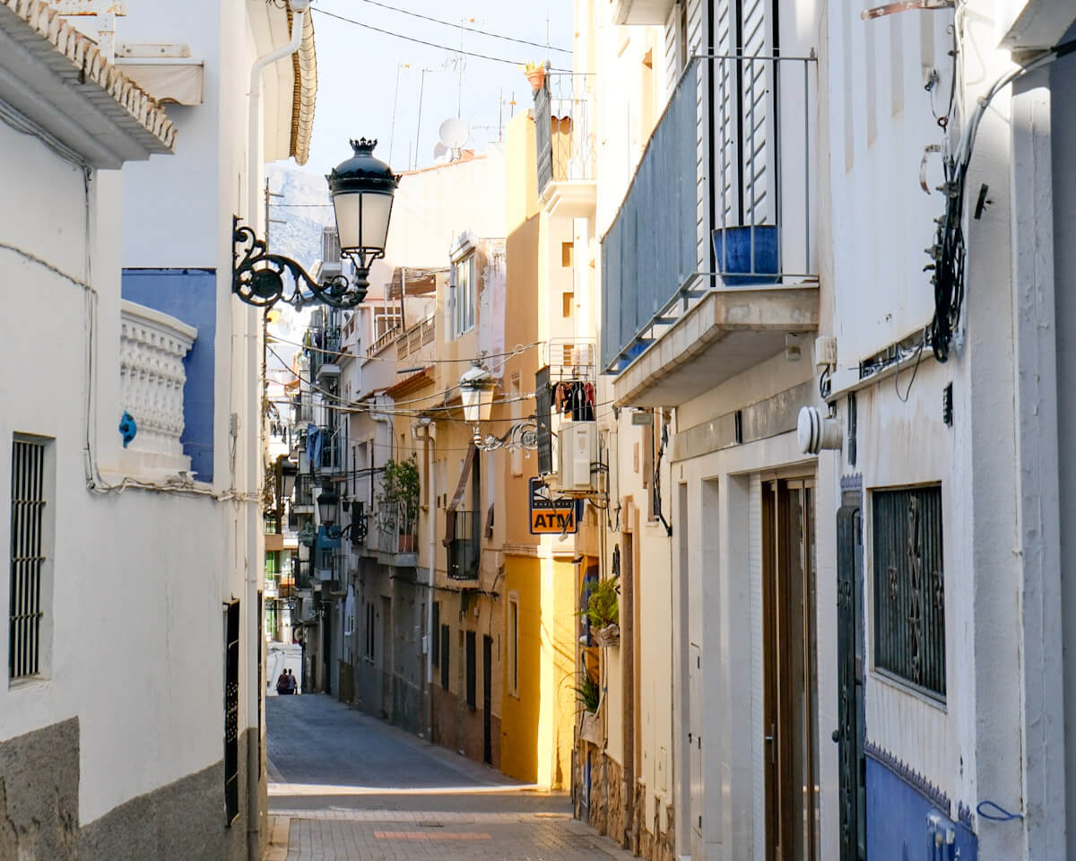 A quiet, sunlit alleyway in historic Benidorm Old Town featuring traditional white and yellow building facades, wrought-iron balconies, and vintage street lanterns