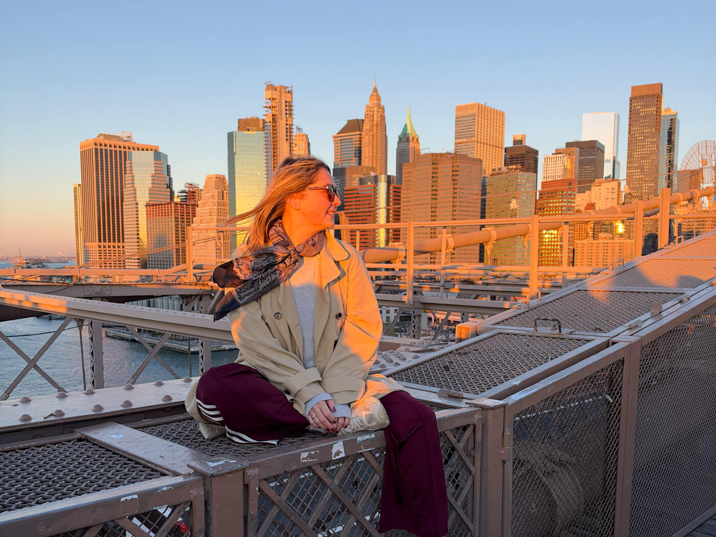 Charlie sitting on the Brooklyn Bridge walkway wearing a trench coat and scarf during a golden sunrise over the Manhattan skyline.