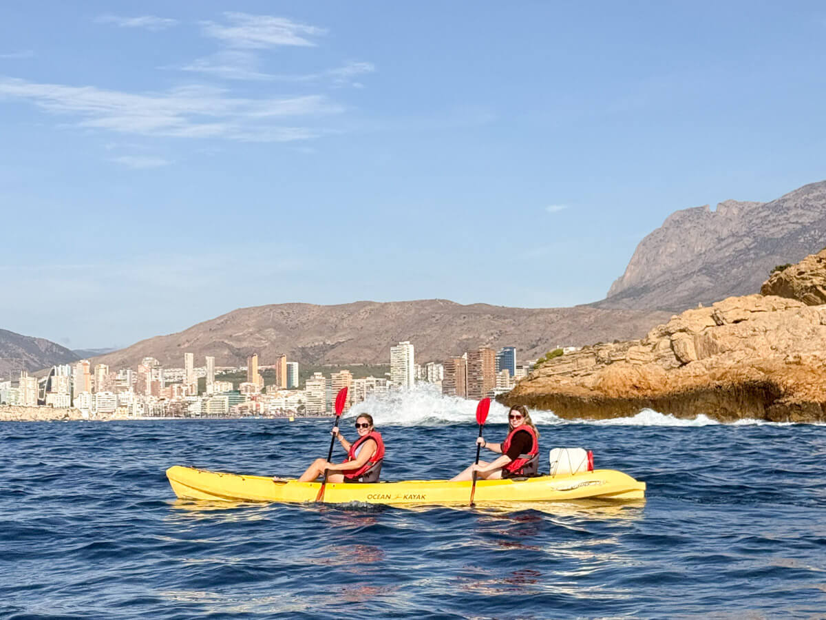 Two women paddling a bright yellow tandem kayak in the blue sea, with the Benidorm city skyline and coastal cliffs in the background, showcasing water activities for what to do in Benidorm without partying.