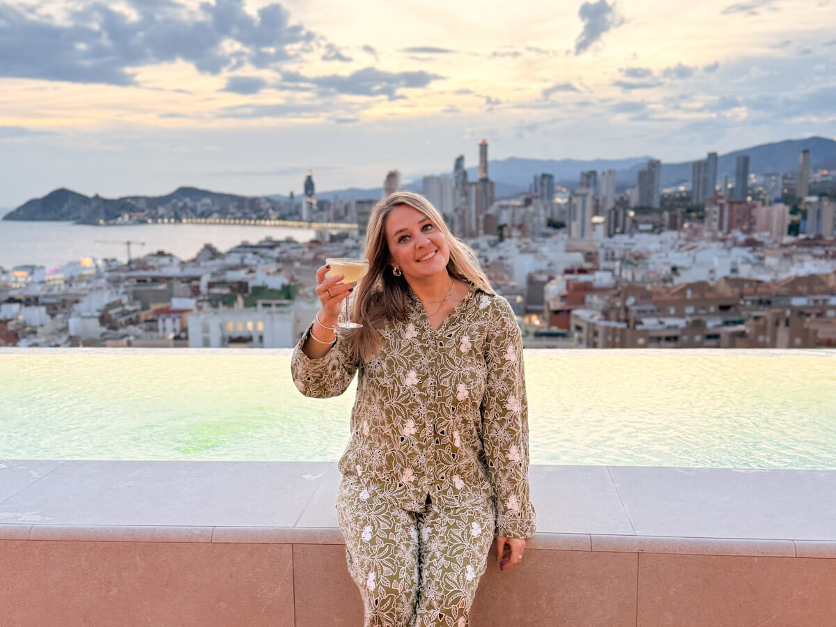 Charlie is smiling while holding a cocktail coupe sitting in front of an infinity pool at Selvatico Rooftop Bar, with panoramic sunset views over the Benidorm coastline and skyline.