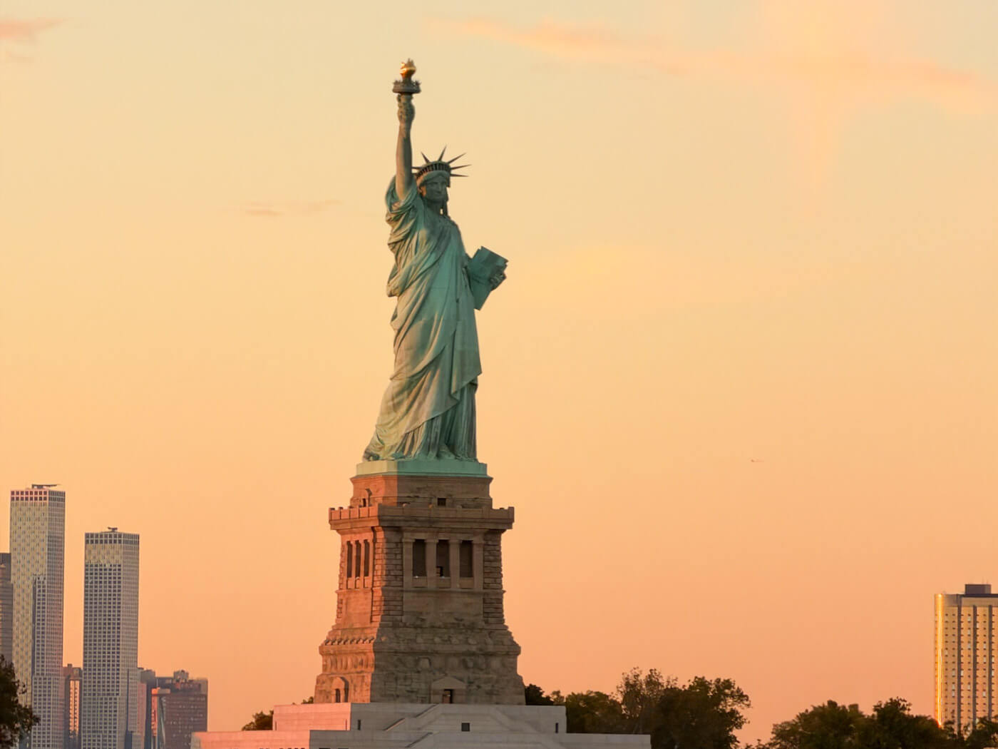 The Statue of Liberty stands illuminated against a warm orange sunset sky in New York Harbor.